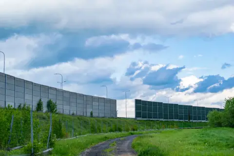Długie ekrany akustyczne drogowe chronią przed hałasem. Betonowe i zielone panele wzdłuż drogi, na tle błękitnego nieba z chmurami.