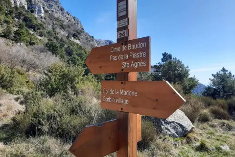 Panneau indicateur de randonnée : direction Cime de Baudon, Pas de la Piastre, Ste-Agnès, Col de la Madone et Gorbio village. Paysage montagneux.