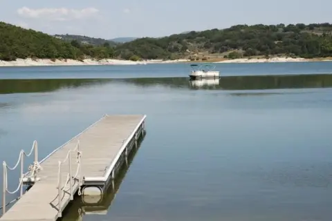 Un pontón blanco navega en el tranquilo embalse de Prada, con un muelle de madera en primer plano y colinas verdes al fondo.