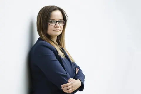 Kobieta w okularach i granatowej marynarce, oparta o ścianę, ze skrzyżowanymi rękami.