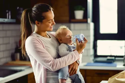 Uśmiechnięta mama karmi butelką niemowlę. Kiedy można podać koperek dla noworodka? To pytanie nurtuje wielu rodziców.