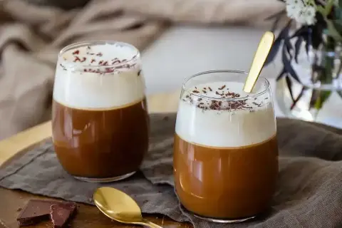 Två glas med krämig kaffe Karlsson, toppad med vispgrädde och chokladströssel. En gyllene sked ligger i ett av glasen.