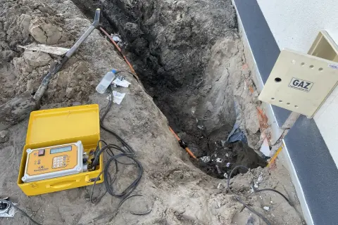 Wykonanie przyłącza gazu od skrzynki do domu. Koparka odsłoniła rurę gazową, obok leży żółta walizka z aparaturą pomiarową.