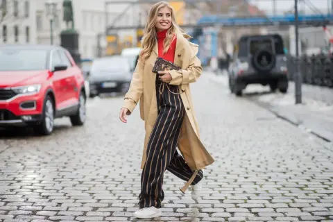 Frau in Beige-Trenchcoat, rotem Rollkragenpullover und gestreiften Hosen, die zeigt, wie man einen Trenchcoat kombinieren kann.