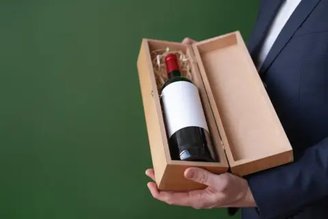 Elegancki mężczyzna w garniturze prezentuje drewniane pudełko z butelką wina wytrawnego czerwonego.