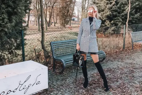 Stylowa kobieta w wełnianym płaszczu w pepitkę, czarnych kozakach i okularach przeciwsłonecznych pozuje w parku.