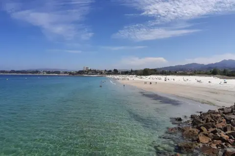Amplia vista de la playa del Vao en Vigo, con aguas cristalinas, arena blanca y gente disfrutando del sol.