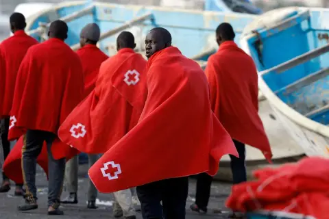 Grupa migrantów w czerwonych kocach z białym krzyżem, po trudnej podróży, szuka schronienia.