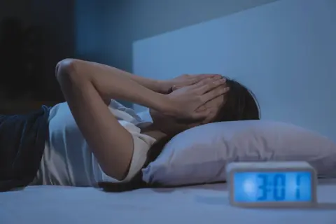 Kobieta w łóżku zakrywa twarz dłońmi, budzik pokazuje 3:01. Brak snu, brak endorfin.