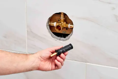Hand holding a shower valve cartridge next to an open wall cavity revealing the shower valve body.