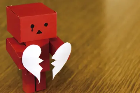 Un robot rojo de origami, con cara triste y una lágrima, sostiene las dos mitades de un corazón blanco. Parece estar a punto de morir de amor.