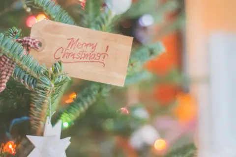 Zawieszka na choince z napisem "Merry Christmas!" w języku angielskim, otoczona igliwiem i migoczącymi światełkami.