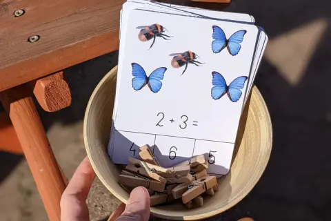 Karty pracy z owadami i zadaniami matematycznymi, np. 2+3=, w miseczce z klamerkami.