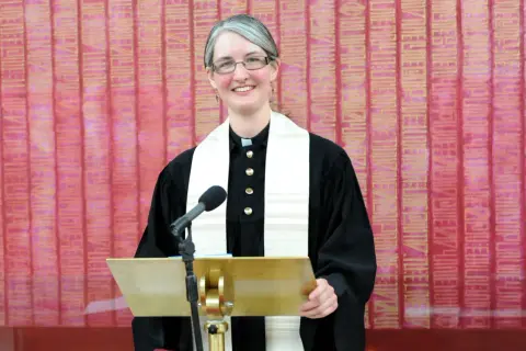 Eine Pfarrerin mit Brille und grauem Haar steht lächelnd hinter einem goldenen Pult. Sie trägt eine schwarze Robe und ein weißes Stola. In dir steckt ein guter Christ.