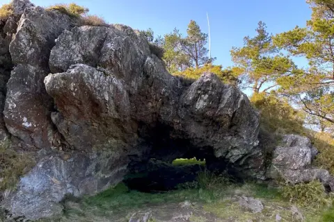 Entrada a la cova da serpe, una cueva natural entre rocas y vegetación bajo un cielo azul.