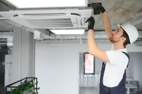 Instalator w kasku montuje klimatyzator kasetonowy na suficie.