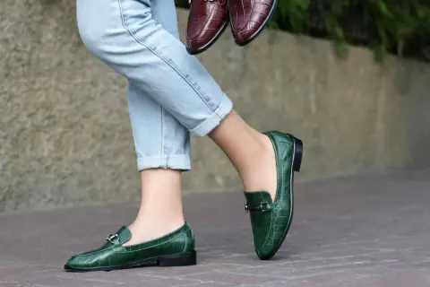 Un paio di jeans chiari e un paio di eleganti mocassini uomo verdi con fibbia metallica. Un altro paio di mocassini marroni è tenuto in mano.