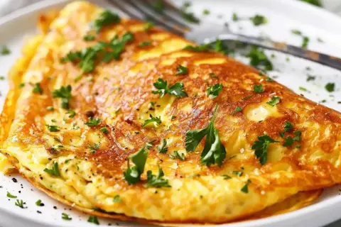 En gyllene omelett, perfekt för ett snabbt omelett recept, toppad med färsk persilja och serverad på en vit tallrik med en gaffel.