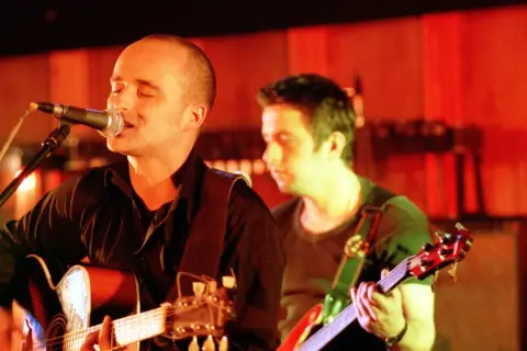 Wokalista z gitarą śpiewa do mikrofonu, i nawet kiedy będę sam, czuje wsparcie kolegi z zespołu.