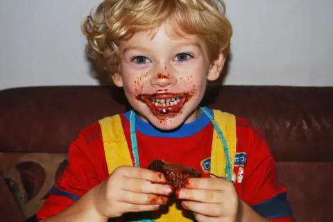 Bambino con cioccolato in bocca e sulle mani, che tiene un pezzo di torta.