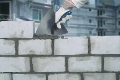 Maurer legt Mörtel mit Kelle auf Ziegelsteine. Der Unterschied zwischen Zement und Beton wird hier beim Mauern deutlich.