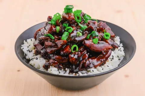 En skål med ris toppad med glansig teriyaki kyckling och strimlad salladslök.