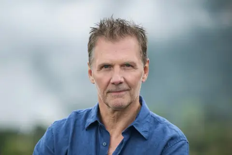 Johan Gullberg, en man med väderbitet ansikte och rufsig frisyr, bär en blå skjorta. En bild som väcker tankar, nästan som en flashback.