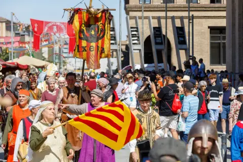 Gente vestida de época desfila en la feria medieval Zaragoza. Un estandarte con un dragón y la palabra "IXERA" ondea.