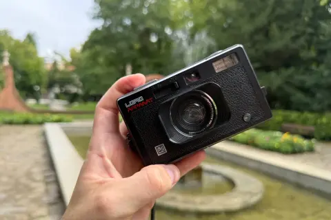 Czarna, analogowa lomografia LomoApparat z obiektywem 21mm. W dłoni, na tle zieleni i fontanny.