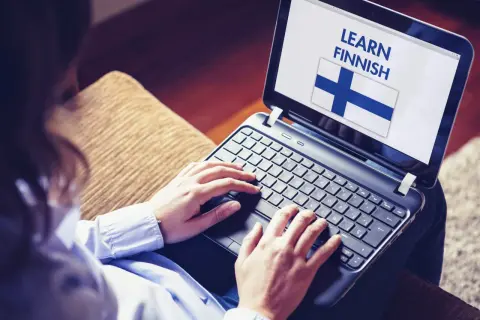 Osoba uczy się języka fińskiego na laptopie, widoczna flaga Finlandii i napis "LEARN FINNISH".