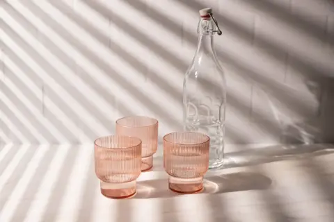 Natural product photography: three pink ribbed glasses filled with water and a clear glass bottle with a swing top, bathed in dappled light.