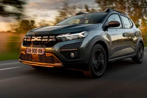 Zielony Dacia Sandero Stepway pędzi po drodze o zachodzie słońca, otoczony rozmazanymi drzewami i mglistym krajobrazem.