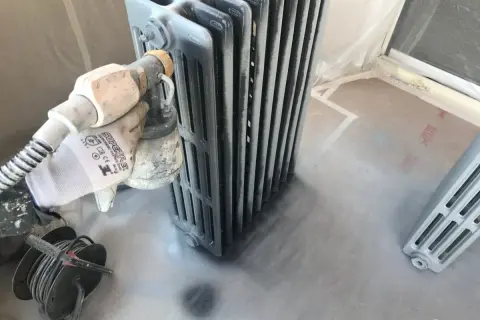 Proceso de pintar radiadores de hierro con pistola. Guante protege mano mientras se aplica pintura gris.