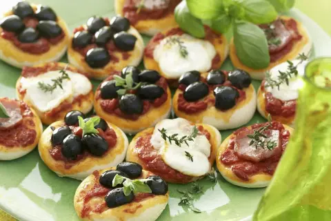 Kleine Pizzen mit Oliven und Mozzarella auf einem grünen Teller, garniert mit Basilikum.