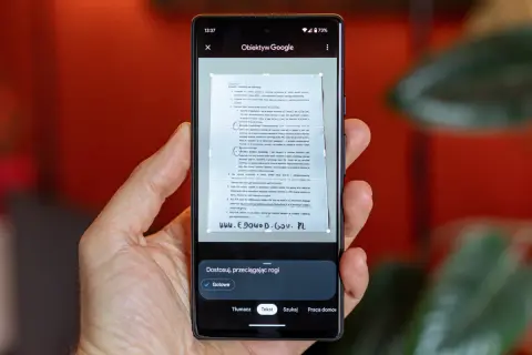 Ręka trzyma smartfon, na ekranie którego działa skaner dokumentów. Aplikacja Obiektyw Google skanuje tekst z papierowego dokumentu.
