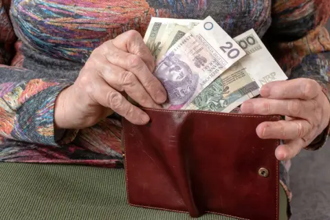 Dłoń starszej osoby wkłada polskie banknoty do brązowego portfela. To pieniądze, ich ekwiwalent.