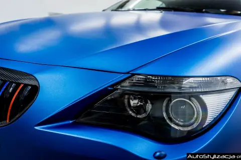 Matowy, **laguna blue** lakier na masce i błotniku sportowego auta. Widoczny fragment reflektora i grill z akcentami M.