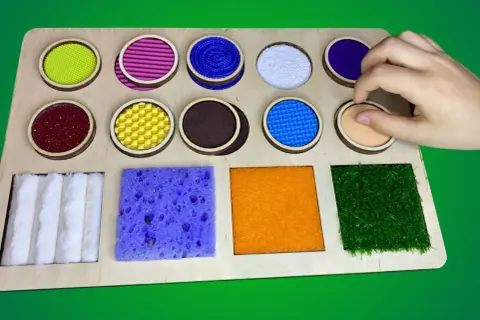Tabla sensorial con diversos tipos de texturas para niños: círculos con materiales variados y cuadrados con esponja, fieltro y césped artificial.
