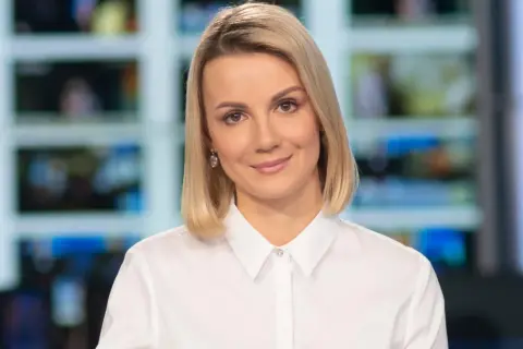Joanna Dunikowska-Paź, znana z Wikipedii, w białej koszuli, z kolczykami, na tle ekranów.