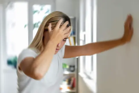 Femme blonde se tenant le front, appuyée contre un mur, le visage pâle, signe d'un malaise vagal.