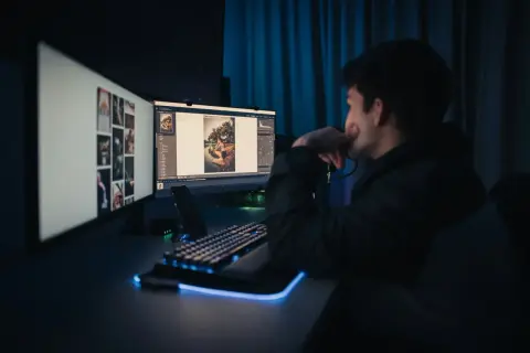 Un jeune homme réfléchit devant son **ordinateur retouche photo**, éclairé par la lueur des écrans.