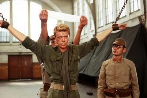 David Bowie jako major Celliers, pojmany w "Merry Christmas Mr. Lawrence", z rękami przykutymi do łańcuchów. Obok stoi japoński oficer.