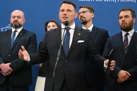Mężczyzna przemawia przy mikrofonie, otoczony innymi politykami. W tle niebieska ściana z napisem. Konfederacja postulaty.