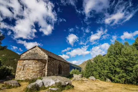 Pequeña ermita de piedra en las rutas del Vall de Boí, rodeada de naturaleza y bajo un cielo azul con nubes algodonosas.