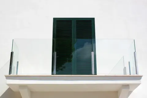 Nowoczesny balkon z przeszkloną balustradą i ciemnozielonymi okiennicami. Okapnik balkonu zapewnia ochronę przed deszczem.