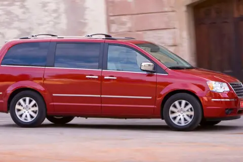 Czerwony Chrysler Grand Voyager w ruchu, z przyciemnianymi szybami i relingami dachowymi, przemierza ulicę.