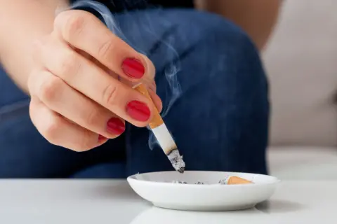 Eine Hand mit roten Nägeln hält eine brennende Zigarette über einem Aschenbecher. Rauch steigt auf und hinterlässt einen kalter rauch geruch.