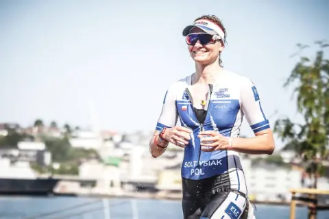 Uśmiechnięta sportsmenka w stroju do triathlonu, biegnie z kubkiem wody. To właśnie triathlon co to za dyscyplina sportu!