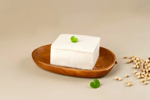 Kostka białego tofu na drewnianej misce, obok nasiona soi. Tofu powstaje z ziaren soi.