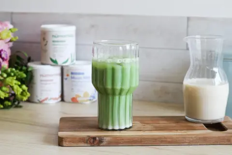 Un verre de matcha latte frais, avec des pots de poudre de matcha et une carafe de lait végétal en arrière-plan.